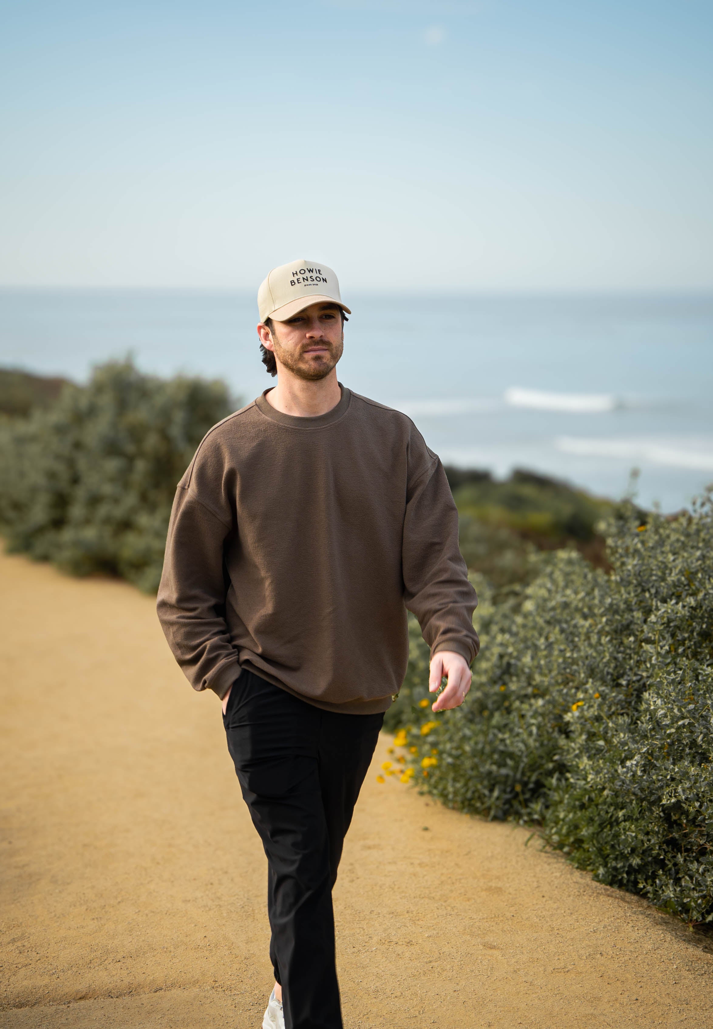 Man walking on a path with ocean view wearing a brown sweatshirt and beige cap.